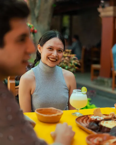 Smiling woman in a gray sleeveless top seated at a table outdoors with food and a cocktail garnished with a lime slice.