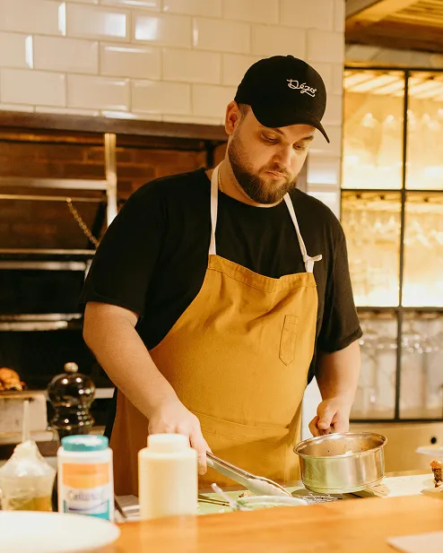 Man wearing a black cap and brown apron cooking with a pan in a kitchen.
