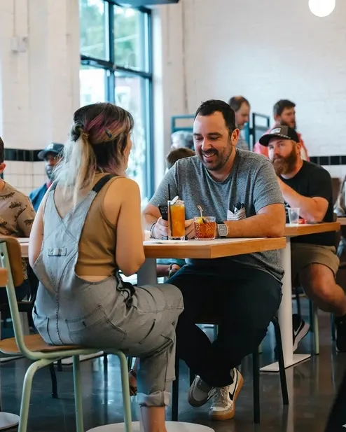 A man and woman sitting at a cafe table enjoying drinks and chatting, with other patrons in the background.